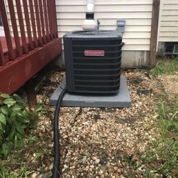 New unit - The image shows an outdoor Goodman air conditioning unit placed on a concrete pad beside a house with beige siding. A drain pipe runs vertically above the unit, and a black power cord is connected to the side of the unit, extending toward the foreground. The ground around the unit is covered with small rocks and some grass and plants. A wooden deck with red-painted railing is visible on the left side of the image.