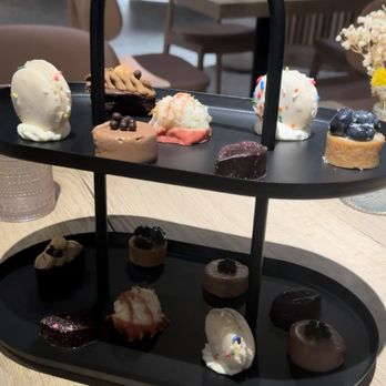 Pesca Vilano - A black, two-tiered dessert stand displays an assortment of small desserts such as cookies, chocolates, and confections. The stand is placed on a light wooden table in a dining area with chairs and tables visible in the background. The desserts are decorated with colorful sprinkles, fruit, and chocolate, and the setting appears to be a restaurant or cafe with a cozy ambiance. - Seafood Near Me - St. Augustine, Florida