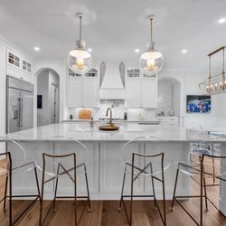 AGC Home Remodeling - Luxury white kitchen with marble island, gold barstools, glass pendants, custom cabinets, and warm wood floors in Lake Austin.