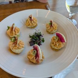 Deviled Eggs - Six deviled eggs with piped filling are decorated with small herb leaves and thin slices of radish. Microgreens are placed in the center of the plate. The dish is set on a wooden table, with a glass of water nearby, and part of a person's arm is visible in the background.