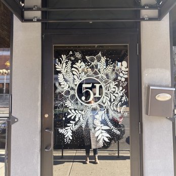 Refinery 51 - Outdoor mural - Glass door with a white botanical design surrounding the number 51, which is centered on the door. The reflection shows a person taking the photo, standing on a sidewalk with a vehicle and buildings visible in the background. The door is framed by a black metal structure, and there is a black canopy above with the number 51 displayed. - Cocktail Bars Near Me - Poughkeepsie, New York