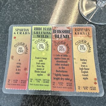 Sowams Cider Works - Four paper strips with cider descriptions and alcohol content are laid out on a dark surface, with a wine glass partially visible. - Cideries Near Me - Warren, Rhode Island