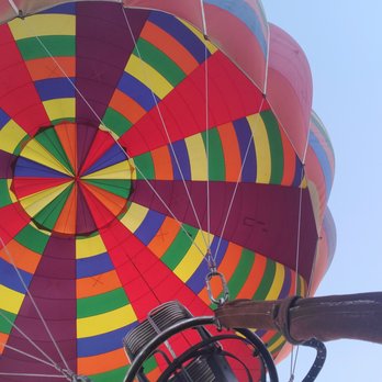 Fly Albuquerque - Fly Albuquerque  flyalbuquerque.com - A vibrant, multicolored hot air balloon is shown from below, with a person in the basket holding onto the edge, against a clear blue sky. - Hot Air Balloons Near Me - Albuquerque, New Mexico