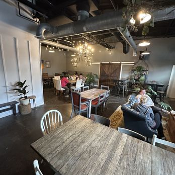 Grutogi Bistro - Back area seating - The image shows the interior of a cafe with a mix of wooden tables and chairs, some with cushioned seats. Several customers are seated, engaged in conversation or using devices. The ceiling has exposed ductwork and industrial-style lighting, including chandeliers and pendant lights. Decor includes plants, artwork on the walls, and a shelving unit with decorative items. The space appears warm and inviting, with a combination of modern and rustic design elements. - Coffee & Tea Near Me - Keller, Texas