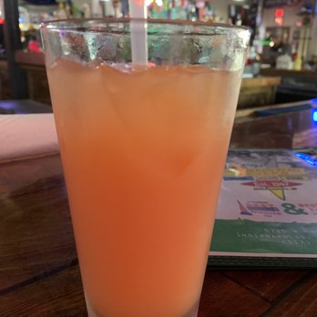 Checkered Flag Tavern - A Hurricane - A tall glass filled with a pinkish-orange beverage and ice, with a straw, placed on a wooden bar table in a lively bar setting. - Pubs Near Me - Indianapolis, Indiana