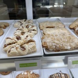 Bakery display case featuring apple blossom pastries with icing and caramel drizzle, and strudel pie with powdered sugar.
