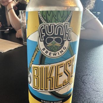 Funk Brewing - A can of Funk Brewing's Lemon Shandy, featuring a colorful label with the Funk Brewing logo and the word 'BIKES,' is positioned on a dark table. The label indicates it is an ale brewed with lemon juice and is always in season. In the background, two women are seated at the table, one with glasses and a bracelet, and the other with curly hair, both engaged in their activities. A glass of water and a napkin are also visible on the table. - Breweries Near Me - Emmaus, Pennsylvania