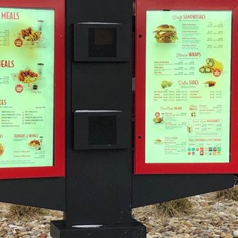 Slim Chickens - Drive-Thru Menu - Two illuminated menu boards at Slim Chickens in Avon, Ohio, display various food options including tender meals, wing meals, house sauces, chicken and waffles, tenders and wings, craft sandwiches, house wraps, sides, salads, sweets, milkshakes, and beverages. The menu features images of food items and prices, with a black background and red framing around the boards. The setting appears to be outdoors, with gravel and some grass visible at the base of the display. - Fast Food Near Me - Avon, Ohio