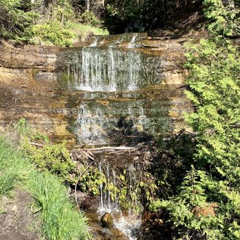 Alger Falls - Alger Falls is scenic in afternoon time. - A waterfall with multiple levels flows over rocks in a forest, surrounded by green trees and vegetation. - Hiking Near Me - Munising, Michigan