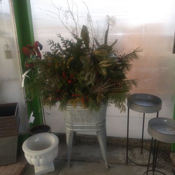 Ken's Gardens - A large floral arrangement featuring a mix of green leaves, branches, and red accents is placed in a metal container on a concrete floor. Nearby, there are decorative items including a flamingo figure, a white plant pot, and two small round metal plant stands. The background includes a translucent wall with green framing. - Nurseries & Gardening Near Me - Lancaster, Pennsylvania