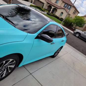 1daywraps.com - A blue car is parked on a concrete driveway in a residential area. The vehicle has tinted windows and black side mirrors. The driveway is adjacent to a street with other parked cars and well-maintained houses with landscaped yards. The sky is partly cloudy, and a streetlamp is visible near the car. The reflection of trees and clouds can be seen on the car's windshield. - Marketing Near Me - Murrieta, California