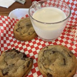 Cookie plate - A glass of milk and three chocolate chip cookies are placed on red checkered paper, with a game board and utensils visible in the background.