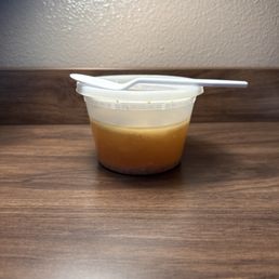 Wonton soup broth. - A clear plastic container filled with broth, topped with a white spoon, sits on a wooden table against a plain wall.