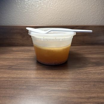Thai House - Wonton soup broth. - A clear plastic container filled with broth, topped with a white spoon, sits on a wooden table against a plain wall. - Thai Near Me - Red Bluff, California