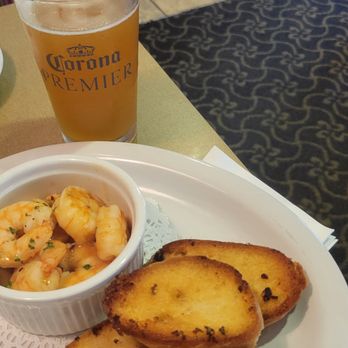 Abingdon Grill - A dining scene with a plate holding cooked shrimp in a small bowl, accompanied by slices of garlic bread on a white plate. A glass of Corona Premier beer is also visible on the table. The table has a beige tablecloth, and the background shows a patterned carpet and part of a chair. - American Near Me - Abingdon, Maryland