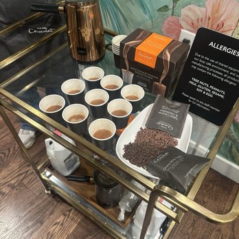 Hotel Chocolat - A gold-colored beverage cart holds cups of hot chocolate, a box of chocolate, chocolate shavings, and a hot water dispenser, with allergy information displayed. - Chocolatiers & Shops Near Me - Chicago, Illinois