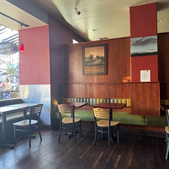 Project Burger - Cool layout with warm colors - The image shows the interior of a restaurant with a combination of wood-paneled walls and painted sections. There are green cushioned benches along the wall with wooden chairs and tables in front. Two landscape paintings are hung on the wooden wall. Natural light from a large window on the left illuminates part of the space, and the ceiling has recessed lighting and a small spotlight. The flooring is dark wood. - American Near Me - Alameda, California