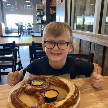 Upland Brewing - Bloomington - Giant Pretzel made with their Beer Cheese and Beer Mustard - A boy with glasses is seated at a restaurant table, smiling at the camera, with a large pretzel and dipping sauces in front of him. - American Near Me - Bloomington, Indiana