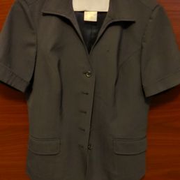 Women's short sleeve button up top - A women's short sleeve blazer in gray fabric with a collar and front buttons, hanging on a hanger attached to a wooden door. The blazer features two front pockets and appears to be made of a structured material suitable for professional or casual wear.