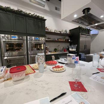 The Recipe - A modern kitchen with dark green cabinets, stainless steel appliances, and open shelves, featuring baking supplies and decorated macarons on the counter. - Venues & Event Spaces Near Me - Ames, Iowa