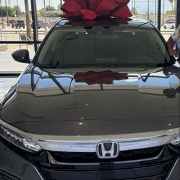 Love my car - A black Honda vehicle with a large red bow on its roof is displayed inside a dealership showroom with large glass windows and a view of the parking lot outside.
