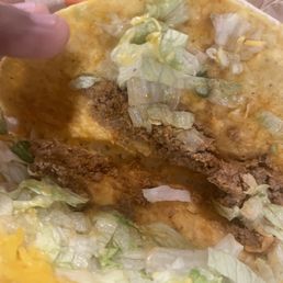 Legit no meat - A taco Bell taco with a soft taco shell contains shredded lettuce, shredded cheese, and fried chicken pieces. The taco is partially eaten, revealing the filling inside. The taco is placed on taco bell branded paper, with some lettuce and cheese visible around it.