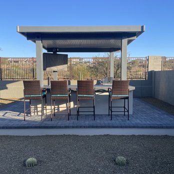 Diamond Cut Landscaping - An outdoor bar area features four wicker chairs positioned at a bar counter, which is situated on a brick-paved patio. The setup is covered by a roof structure, with a TV mounted on the back wall. The surrounding area includes a cacti plant and a gravel ground, enclosed by a concrete block wall and metal fencing, with a clear blue sky overhead. - Landscaping Near Me - Phoenix, Arizona