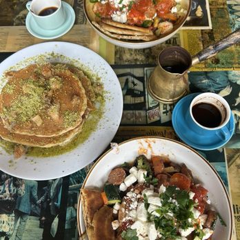 Brookline Lunch - Baklava pancakes and shakshuka - A table features three dishes: a plate of baklava pancakes topped with pistachios and syrup, a bowl of spicy sausage shakshuka with vegetables and poached eggs, and a salad with cheese, vegetables, and herbs. Two cups of black coffee are present, along with a cezve for brewing coffee. The table surface has a newspaper print design, and the setting suggests a casual dining environment with Middle Eastern and American fusion flavors. - Diners Near Me - Cambridge, Massachusetts