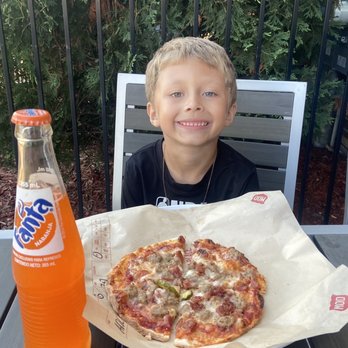 MOD Pizza - Kids pizza with a Fanta - A young boy with short light brown hair and a black shirt is seated at an outdoor table, smiling at the camera. In front of him is a pizza on a paper-lined tray and a bottle of Fanta orange soda. The background features green foliage and a black metal fence, indicating an outdoor dining area. - Fast Food Near Me - Rockford, Illinois