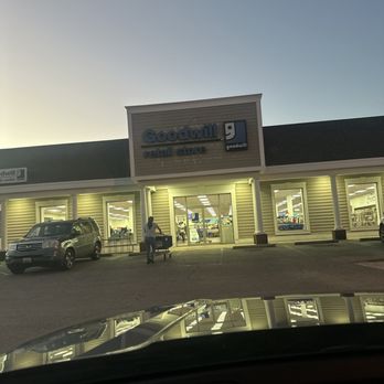 Goodwill - Never a good selection - The image depicts the front exterior of a Goodwill retail store during dusk or early evening. The store has large, illuminated windows and a sign displaying the Goodwill logo and name. There are parked vehicles in the parking lot, and a person is seen pushing a shopping cart near the entrance. The photo appears to be taken from inside a vehicle, with the dashboard visible at the bottom of the frame. - Thrift Stores Near Me - Waldorf, Maryland