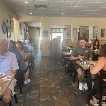 The Pantry - West view of restaurant from south east corner of restaurant - The image shows the interior of a restaurant with several customers seated at tables, some engaged with their phones and others eating or conversing. The space has a warm, inviting atmosphere with wall decorations and large windows allowing natural light. A staff member is visible in the background, attending to the dining area. The flooring is tiled, and the ceiling has recessed lighting. - Diners Near Me - Westerly, Rhode Island