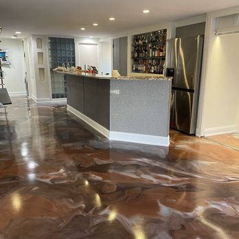 AD Technology - Epoxy flooring in my family room. - The image displays a polished epoxy floor with a marbled, swirling pattern in shades of brown, beige, and cream. The floor reflects light, indicating a high-gloss finish. In the background, there is a bar counter with a textured surface, a stainless steel refrigerator, and a modern kitchen area with white walls and recessed lighting. The space appears to be part of a residential interior designed for entertaining. - Flooring Near Me