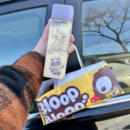 A person holds a milk bottle with panda-themed illustrations and a purple cap, alongside a yellow snack bag with a cartoon face, outside near a black vehicle.