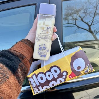Bloop Bloop - A person holds a milk bottle with panda-themed illustrations and a purple cap, alongside a yellow snack bag with a cartoon face, outside near a black vehicle. - Sushi Bars Near Me - Chicago, Illinois