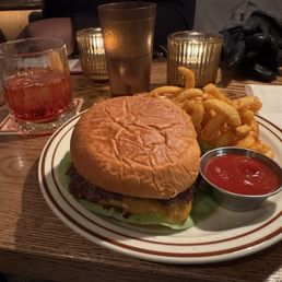 Smash Burger and Mezcal Negroni - A meal featuring a cheeseburger with a soft bun, lettuce, and a beef patty, accompanied by curly fries and a small metal cup of ketchup, is presented on a striped ceramic plate. The setting includes a wooden table with drinks, including a glass of red beverage, a glass of water, and a frosted glass of a cold drink, with candles and other tableware in the background.