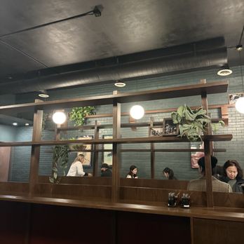 Dumpling Wei - The image shows the interior of a restaurant with a dark ceiling and exposed ductwork. Wooden dividers separate the dining area, with patrons seated at tables along a brick wall decorated with framed photos and potted plants. Hanging globe lights provide illumination, and the overall atmosphere appears modern and cozy. - Chinese Near Me - Pasadena, California
