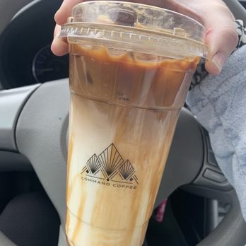 Command Coffee - Iced caramel macchiato - A person holds a large iced caramel macchiato in a clear plastic cup with a lid, featuring the Command Coffee logo. The drink has visible layers of milk, caramel, and coffee. The background shows the interior of a vehicle, including a steering wheel and dashboard. The person's hand and part of their sleeve are visible. - Coffee & Tea Near Me - Indianapolis, Indiana