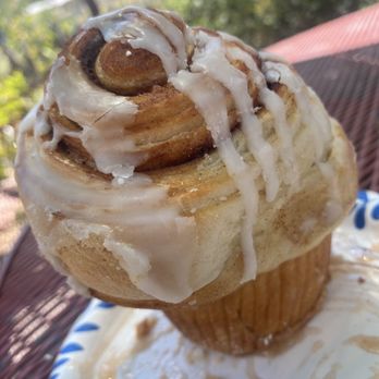 Java Junction - Cinnamon roll - A cinnamon roll with white icing drizzled on top is shown resting on a paper plate outdoors. The background features trees and a red roof, indicating an outdoor setting. The cinnamon roll appears freshly baked with a golden-brown exterior and visible cinnamon swirls inside. - Bed & Breakfast Near Me - Madrid, New Mexico