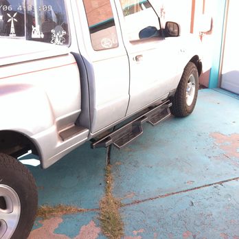 Auto Depot - A silver pickup truck is parked on a blue concrete surface with visible cracks and patches of grass growing through. The truck features step bars along the side, a 4x4 badge near the front wheel, and a side mirror. The background includes a white wall and a partially visible door, with sunlight casting shadows on the ground. - Auto Detailing Near Me - Albuquerque, New Mexico
