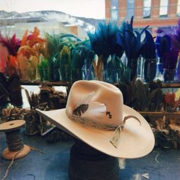 A white cowboy hat featuring a feather and decorative band is positioned on a black hat block on a table. Behind the hat, a window display showcases an array of colorful feathers arranged in glass containers. The background reveals a cityscape with buildings and snow-covered mountains outside the window, indicating a winter setting. The image includes a timestamp and a small icon at the bottom.