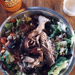 Chopped salad with pulled pork - A chopped salad featuring shredded pulled pork, blue cheese crumbles, and mixed vegetables such as lettuce, tomatoes, and carrots is served in a metal bowl on a wooden table. A bottle of Mango Lemonade and a cup of dressing are also visible in the image, indicating a casual dining setting.