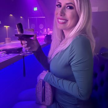 Elysium Immersive Dining - A woman with long blonde hair, dressed in a tight-fitting green dress, is sitting at a bar in a dimly lit venue with purple and blue lighting. She is smiling and holding a cocktail glass with a straw, with her other hand resting on her lap. The background shows a bar counter with some bar equipment and a neon exit sign. The setting appears to be a nightclub or lounge with a modern, stylish atmosphere. - Modern European Near Me - Lighthouse Point, Florida