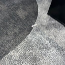 This is a used Covid test swab on the floor by the pharmacy counter - The image shows a used Covid test swab on a gray textured carpet. The swab is small, with a white handle and a pink tip, positioned near the center of the image. A shadow is cast on the carpet, likely from an object outside the frame. A black object, possibly a piece of electronic equipment, is partially visible in the upper right corner. The scene appears to be indoors, on a carpeted floor.
