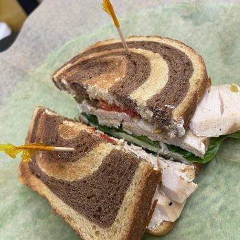 Mercantile - Trendy Farmer - Grilled Chicken Breast with Roasted Red Peppers, Baby Spinach and Merc-made Artichoke Asiago cheese Spread - A marble rye bread sandwich filled with turkey, lettuce, tomato, and cheese, served on green parchment paper with toothpicks. - Bakeries Near Me - Dennis, Massachusetts