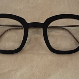 A pair of black eyeglasses with a thick, rounded frame and clear lenses are positioned on a light-colored surface. The glasses have metal temples that extend from the frame, with transparent tips. The image focuses on the glasses, showing their design and construction clearly.