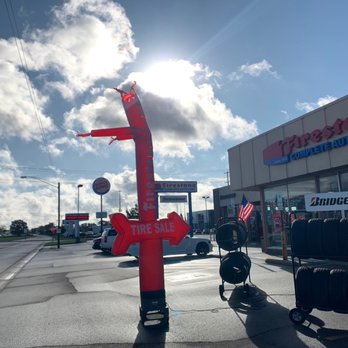 Firestone Complete Auto Care - Front parking lot. - A large inflatable sign shaped like a tire with a red arrow and the words 'TIRE SALE' is situated in the parking lot outside a Firestone Complete Auto Care store. The sign is tethered to the ground, and nearby are stacks of tires and a cart. The store's exterior features signage for Bridgestone tires, and the sky is partly cloudy with the sun shining behind the sign. - Tires Near Me - Appleton, Wisconsin