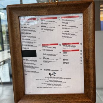 Kohi Time - Current menu... I recommend trying everything! - A wooden-framed menu board from Kohi Time Coffee Counter features a list of coffee beverages, including cold brew, hot coffee, espresso drinks, and specialty options. The menu includes descriptions, sizes, and prices, with sections highlighted in red for hot and cold drinks. The board also notes the business's focus on eco-friendly practices and serving 100% tipless coffee. The background shows a modern cafe interior with large windows and seating areas. - Coffee & Tea Near Me - Eugene, Oregon