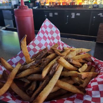 Third Base - Huge portion of fries - A basket filled with French fries is placed on a bar counter, lined with red and white checkered paper. A ketchup bottle is positioned behind the fries. The background shows a bar with illuminated bottles and a blurred television screen, indicating an indoor dining or bar environment. - Dive Bars Near Me - Chandler, Arizona