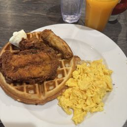 Chicken and Waffle Breakfast - A breakfast plate featuring fried chicken on a waffle, scrambled eggs, a chicken drumstick, and a dollop of butter, with a glass of orange juice and water in the background.