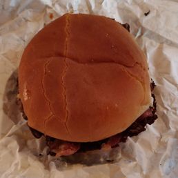 Cowboy Burger - A burger features a soft, toasted bun with visible cracks on the top, placed on crumpled white paper. The burger contains meat and toppings that are partially visible beneath the bun.